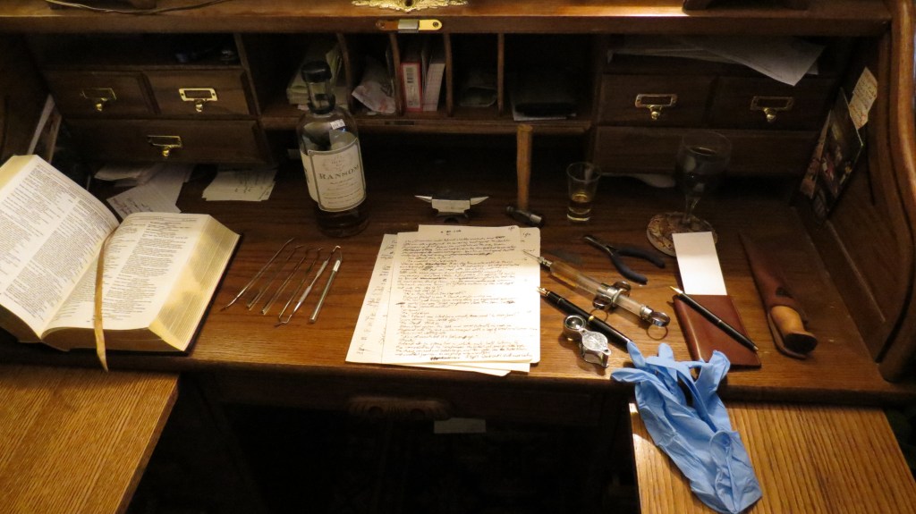 Picture title: Editing. Picture shows the writing surface of a wooden roll-top desk with brass appointments, and the desktop hutch, with three drawers on each side (two small over one large), a "cubby" space over each set of drawers, and five cubby/mail slots over the desk top. On the desk surface, there is a large hard-bound book, with glossy gold edging  on the white pages, and small print. The book is open to a page less than halfway through with an attached ribbon marker across one page. One cover is flat on the desk, and the other is propped against the side-wall where the roll-top would slide down. To the right of the book is a set of five medical or scientific-looking metal tools. They are hooked or bent at one or both ends, possibly from a dissection kit. There is also an exacto knife. Further back from these tools is a bottle of gin with an antiquated label. To the right of the bottle is a tiny jeweler's anvil, with a tiny ballpein hammer next to it. Closer to the camera, on the writing surface of the desk (just to the right of the tools) is a stack of papers with messy handwriting on them. To the right of the papers, arranged facing diagonal to them, is a hand lens, a black fountain pen with a gold nib, an antique glass and steel syringe with an amber fluid in it (it's the gin - just there for show), and a pair of needle nose pliers and a razor blade. Above the pliers is a shot glass with a little more gin. To the right of all that is a wine glass with a black liquid in it, on a stone coaster that shows an old-fashioned map of the world. Below that on the desk is a notepad with a leather case, and another black and gold fountain pen. Next to that is a leather-sheathed knife with a wooden handle. To the right and left, there are wooden pull-out elbow rests. The one on the right has a couple blue surgical gloves on it. The lighting is warm. The overall impression is that to edit the chapter on the papers, the writer will have to perform surgery using the tools on the desk. 